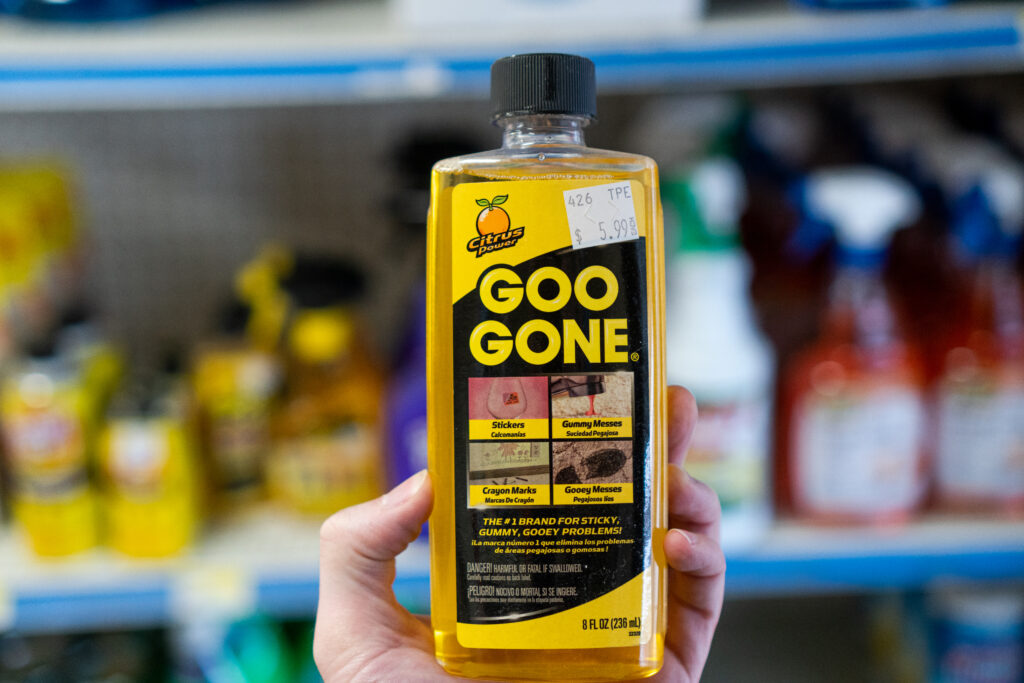 A hand holding a bottle of Goo Gone cleaner in front of a store shelf full of other cleaning products.